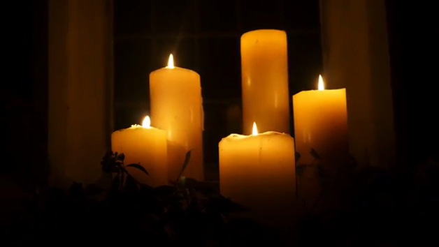 Five lit pillar candles of varying heights glowing warmly in a dark room near a window, with sprigs of greenery